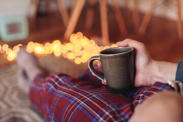 Man holding a cup of tea, wearing check pajamas, sitting at home in a cosy room