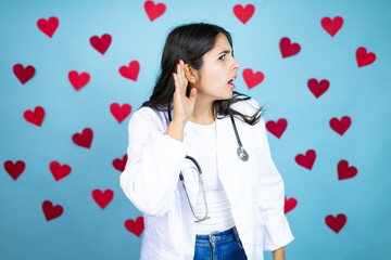 Young doctor woman wearing medical coat and stethoscope over blue background with red hearts surprised with hand over ear listening an hearing to rumor or gossip