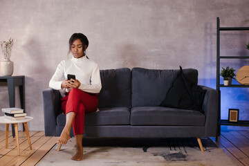 Obraz premium African girl looks at her phone on the sofa in her cozy room