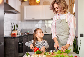 jolly child girl preparing a salad with her mother in the modern light kitchen, enjoy the process of preparing meal