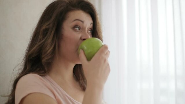 Side portrait of the attractive obese woman eating the green apple with grimacing face on a diet.