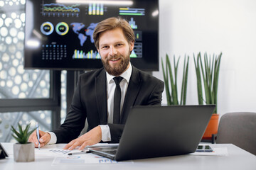 Horizontal indoor shot of young handsome male office worker, sitting at the desk and smiling to camera, while working with laptop pc and making notes. Wall plasma screen on the background