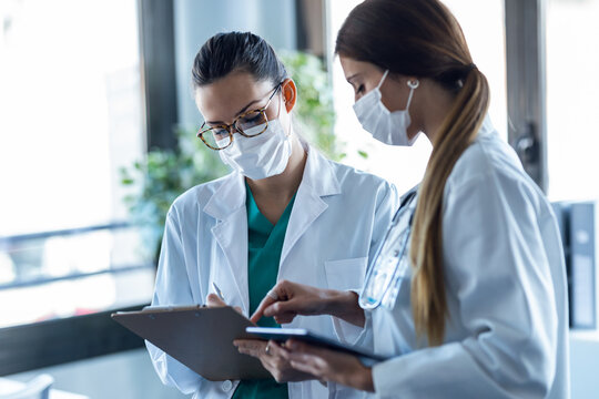 Two Young Women Doctor Wearing A Hygienic Facial Mask Talking With Her Colleague While Working Digital Tablet In Hospital.