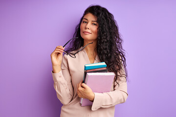 stylish teacher in spectacles posing at camera holding books in hands isolated over purple background