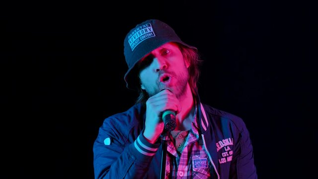 Charismatic musician sings into a microphone in a dark studio on a black background in the light of neon lights. The young guy performs reggae music and moves in a fiery rhythm. Slow motion. Close up.