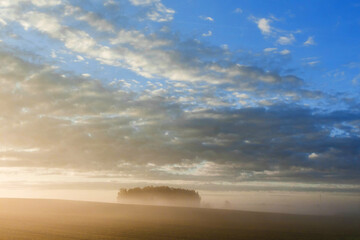 Naklejka na ściany i meble Dawn in a field with the light of the sun from beyond the horizon with blue sky and clouds