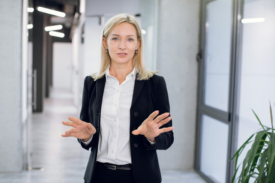 Close Up Portrait Of Mature Blond Caucasian Businesslady, Wearing Formal Suit, Posing To Camera, Speaking And Gesturing With Her Hands, While Standing In Modern Office Corridor Indoors