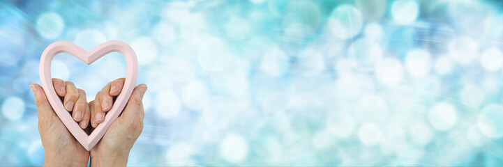 Loving Kindness Heart-centred Giving Campaign banner - female hands holding a pink heart frame against a wide pastel blue bokeh background with copy space for messages