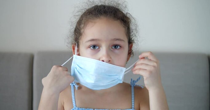 Child looks at the camera and takes off the medical mask. Portrait kid or little girl sitting at home on the sofa wearing a protective medical mask with a sad disappointed face. Pandemic, covid - 19.