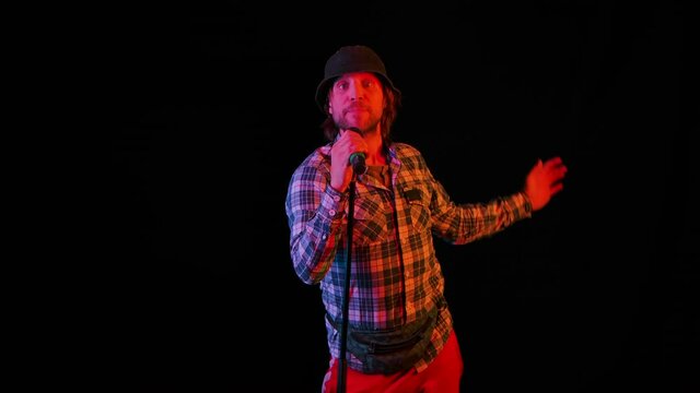 The vocalist's solo performance in a dark studio on a black background in the light of neon lights. The young guy performs reggae music and moves in a fiery rhythm. Slow motion. Close up
