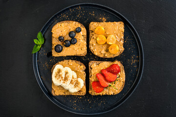 Healthy breakfast toasts with peanut butter and fresh fruits, bananas, strawberries, 
cranberries and physalis on black slate background from top view.