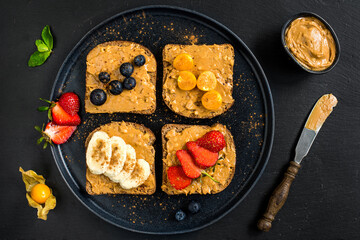 Healthy breakfast toasts with peanut butter and fresh fruits, bananas, strawberries, 
cranberries and physalis on black slate background from top view.