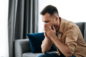 Unhealthy young man. Caucasian guy is sick with flu, he sneezes and blows his nose in a napkin while sitting on the couch at home