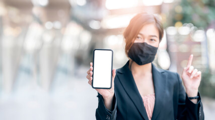 Young asian woman with protective mask showing blank screen smartphone while standing outdoors in the city.