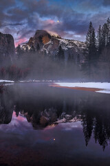 Moon rises above Half Dome in snowy Yosemite National Park at sunset as seen from the Merced river.