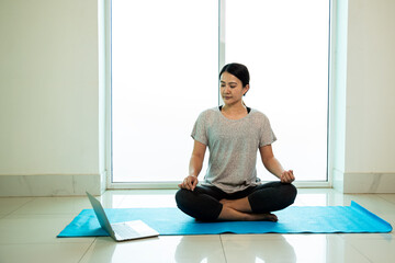 Asian young woman sitting and practices yoga pose on blue mat near window of gym with copy space