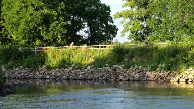A river and trees and a wood fence and two people riding bicycles on a bike path in evening light.