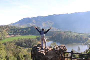 Young man standing on a rock of a cliff and enjoying the view of nature of lake and mountain in Dieng Indonesia