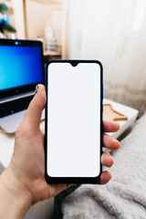 smartphone with white screen in hand on background of workplace in minimalist scandi style with laptop, space for text, vertical content, selective focus