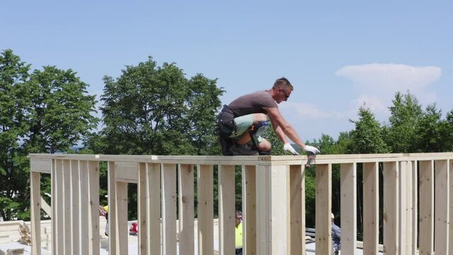 Aerial view of a builder sitting on the unfinished wall and nailing wooden planks. Frame house under construction. Summer