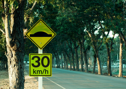 Traffic sign yellow and black, beware of rough roads, do not drive more than 30 kilometers per hour.The backdrop is a road with mahogany trees along the way.