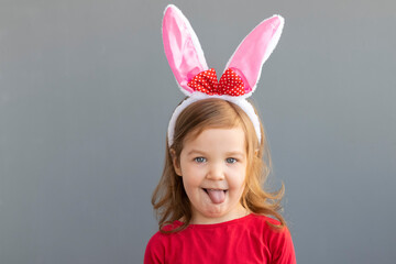 Beautiful little curly red-haired girl with rabbit ears in a red dress on a gray background shows her tongue. Easter holiday concept