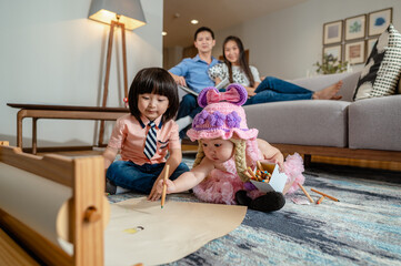 Brother is drawing on paper with little sister on the rug in the living room at home, Parents background sitting on the sofa watching over a distance. Family in home concept.