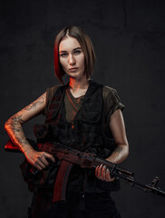 Dressed in black armour with dark shirt martial woman with short haircut poses in dark background holding assault rifle.