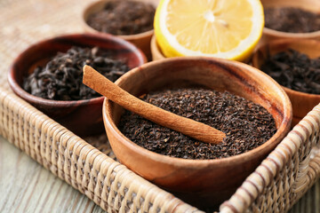 Bowls with dry black tea leaves, cinnamon and lemon on wooden background