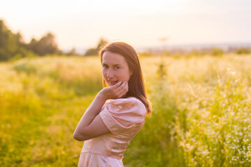 Naklejka na ściany i meble Beautiful portret of happy woman with dark hair standing in the wildflowers field in the long pink dress and sensually touching her face on a warm summer evening. Joyfull summer walking. 