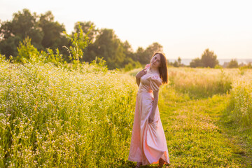 Fototapeta premium Beautiful photo of woman with dark hair standing in the wildflowers field barefoot holding the bottom of the long pink dress on a warm summer evening. Joyfull summer walking. 