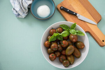 Gestreifte bunte Tiger Tomaten in Schale mit Mozzarella und Basilikum mit Geschirrtuch, Messer, Holz Brett auf Hintergrund Mint Grün
