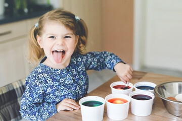 Excited little toddler girl coloring eggs for Easter. Cute happy child looking surprised at colorful colored eggs, celebrating holiday with family. Adorable kid at home with different bright colors.