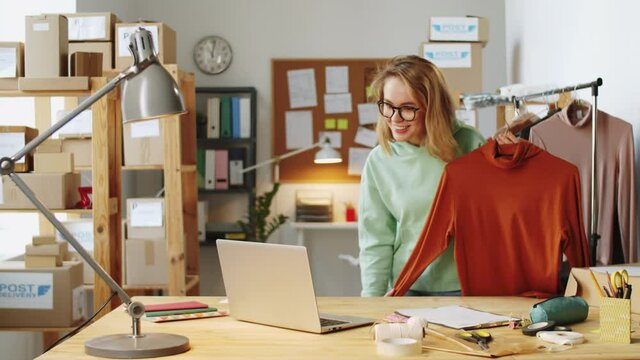Young blonde woman showing and telling about polo neck sweater via online video call on laptop while working remotely with customer in office of online clothes shop