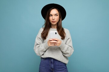 Beautiful young brunette woman thinking wearing black hat and grey sweater holding smartphone looking to the side texting isolated on background