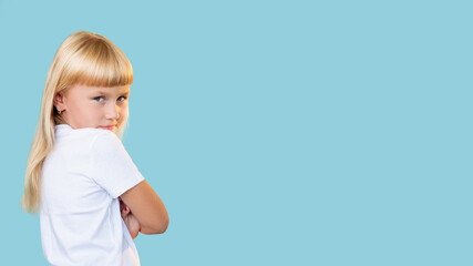 Naughty child. Kids behavior. Education problem. Portrait of offended dissatisfied blonde little girl in white t-shirt looking back with folded arms isolated on blue copy space background.