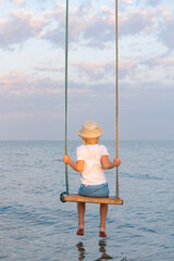 Child is sitting on rope swing over water. Vertical frame. Sea vacation with children