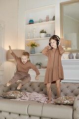 Brother and sister have fun at home and jump on the couch. Children dance and listen to music with headphones