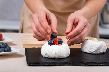Obraz na płótnie Canvas Female hands decorating meringue with blueberries and strawberry. Process of making cakes