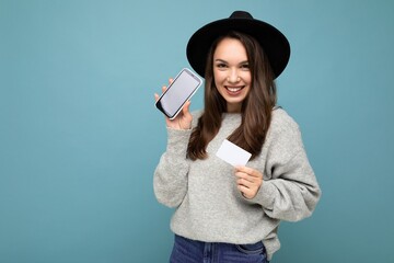 Beautiful smiling young brunette female person wearing black hat and grey sweater isolated over blue background holding credit card and mobile phone with empty display for mockup looking at camera