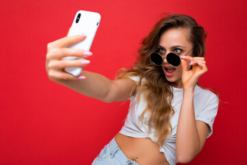 Closeup of happy amazing beautiful young woman holding mobile phone taking selfie photo using smartphone camera wearing everyday stylish outfit isolated over colorful wall background looking at device