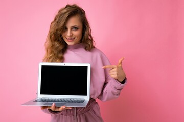 Fototapeta premium Photo of beautiful blond curly young woman lookind at camera holding computer laptop with empty monitor screen with mock up and copy space pointing at netbook with hand and one finger wearing pink