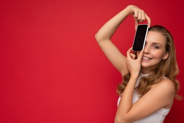 Naklejka premium Photo of beautiful smiling young woman good looking wearing casual stylish outfit standing isolated on background with copy space holding smartphone showing phone in hand with empty screen display for