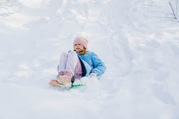 A teen girl in a pink hat and a blue coat tries to move on a green sledge from a snow slide, but the snow is a lot and it does not slide. winter children's walks and fun. clothes for frosts.