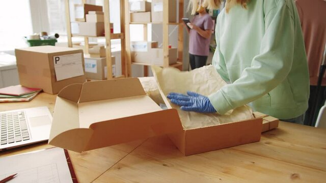 Young blonde woman in protective mask and gloves packing order into box before shipping it to customer while working in office of online store during covid-19 pandemic