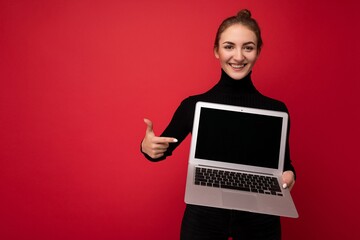 Photo of Beautiful smiling happy young brunette woman holding computer laptop with empty monitor screen wearing black longsleeve looking at camera pinting at netbook isolated over red wall background