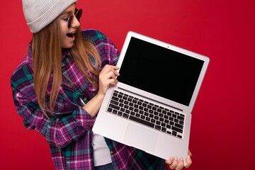 Portrait Photo of beautiful laughing funny smiling blond young woman holding computer laptop with empty monitor screen wearing sun glasses hat and colourful shirt looking at netbook display and