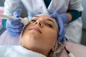 Fototapeta premium young woman receiving professional procedure facial skin ultrasound cavitation from cosmetologist