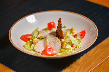 Bowl of Caesar Salad served on a black rustic wooden table in restaurant or diner. Traditional Caesar recipe