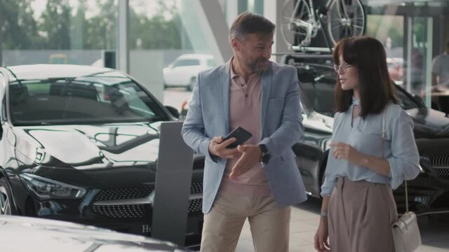 Slowmo shot of cheerful middle-aged man holding mobile phone and taking photo of car in dealership, then talking to his young wife